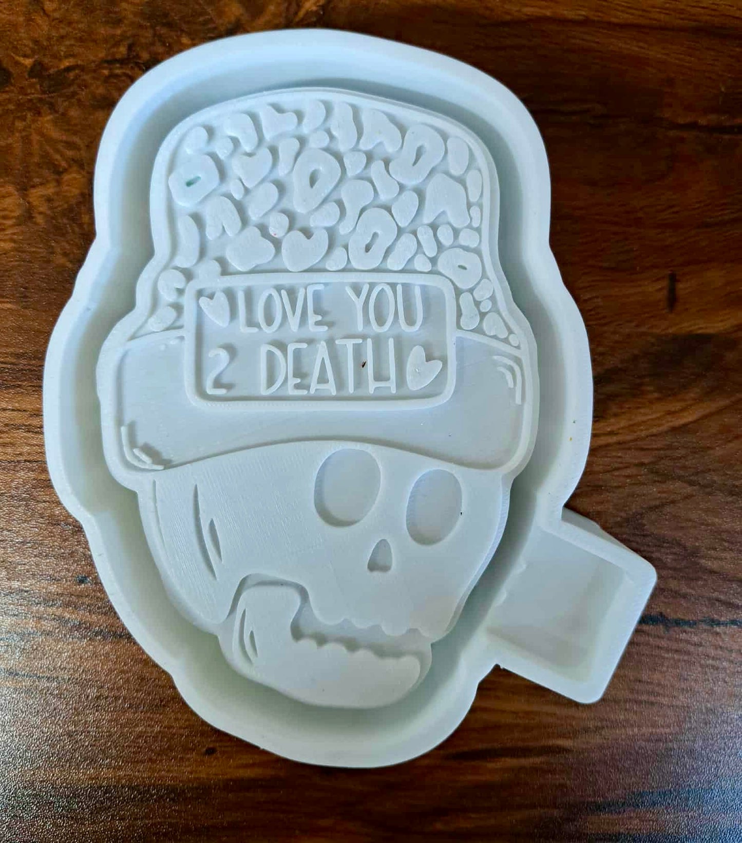 3D Housing shaped like a skull with a cap and text 'I Love You 2 Death' on a wooden surface to make silicone molds. 